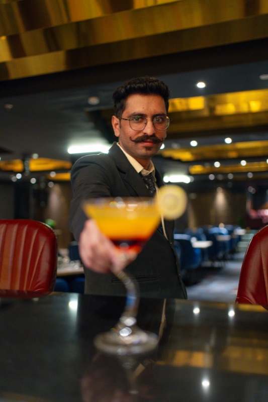 A Bellers server in a suit holding out a layered orange cocktail, the dining room softly lit behind