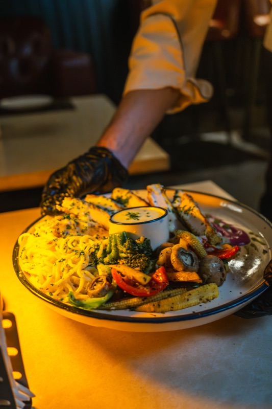 A gloved hand setting down a platter of pasta, grilled chicken and vegetables under a warm light
