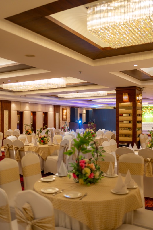 A banquet hall set for an event: round tables in gold and white, a floral centrepiece in front, chandeliers above
