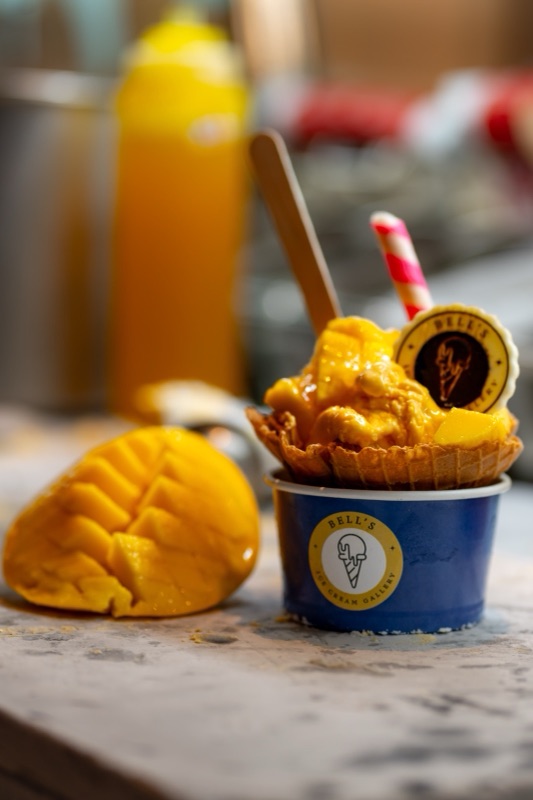 Mango ice cream in a waffle bowl, in a navy Bell’s cup, with fresh mango beside it