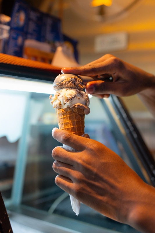 Hands stacking a second scoop onto a waffle cone over the lit freezer