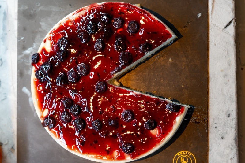 A whole blueberry cheesecake with one slice taken out, from above, on a dark board