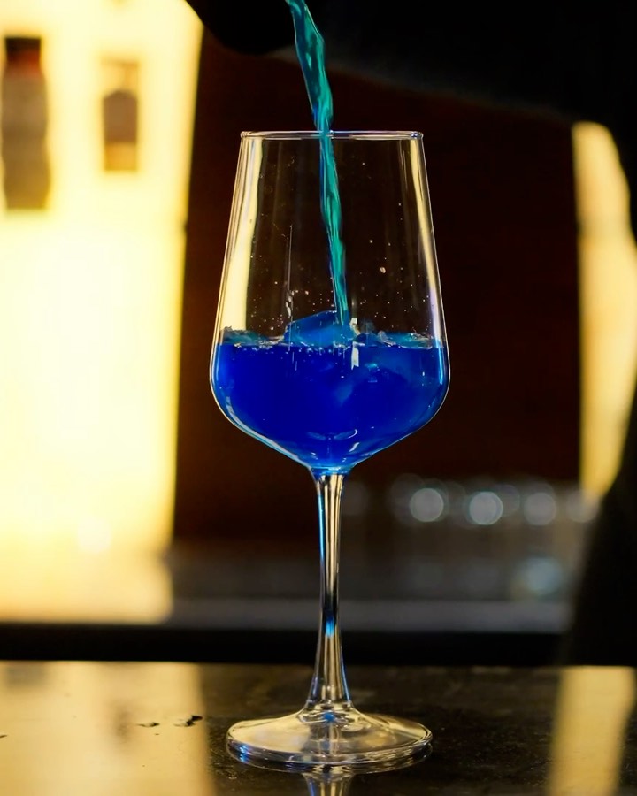 A bartender pours a blue drink over ice into a wine glass at the Bellers bar, the lit bottle wall behind