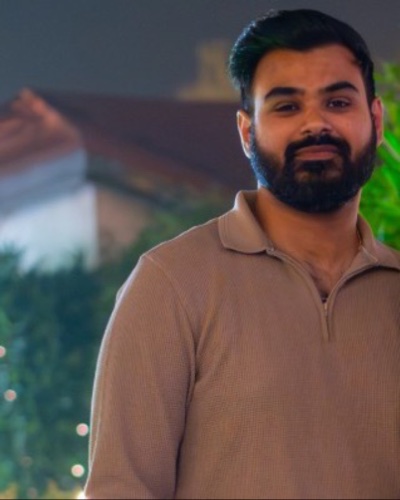 Aditya Vashistha, bearded, in a beige knit polo, in a garden strung with small lights at night