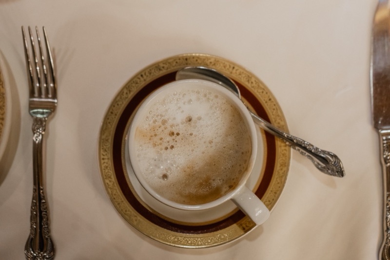 A cappuccino in a gold-rimmed cup on a white tablecloth, from above, with a fork and knife either side