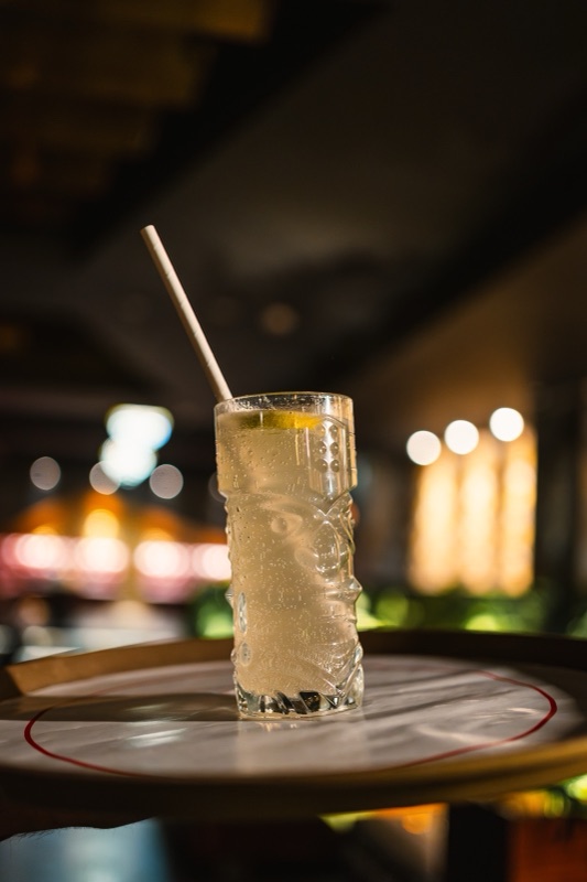 A tall glass of lemon cooler with a paper straw on a round bar tray, warm lights behind